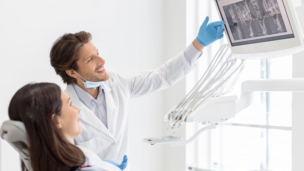 Cheerful male dentist showing a patient their dental X-ray on an LCD screen, discussing treatment planning for full mouth rehabilitation and restorative dentistry solutions.