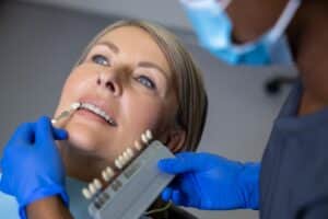 Female dentist holding a dental shade guide to match enamel color for a patient, selecting the right tone to fix unnatural-looking veneers and ensure a natural smile appearance.