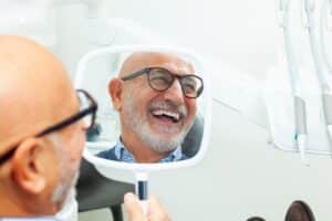 Smiling senior man holding mirror at dental clinic, admiring results of his dental implant treatment.
